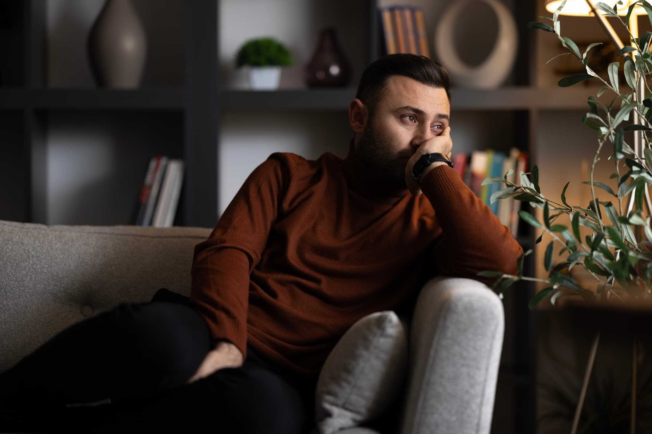 Man med bekymrad min som sitter ensam i soffan – symboliserar psykiska och fysiska orsaker till erektionsproblem. Få snabb hjälp hos Doktera online.