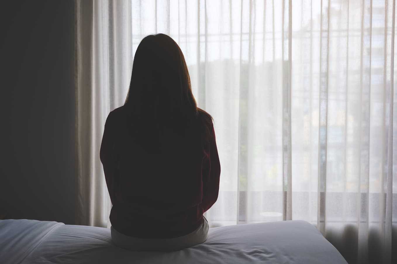 Silhuett av en kvinna som sitter ensam på en säng och tittar ut genom ett fönster – en symbolisk bild av depression. Doktera.se erbjuder läkarbedömning online för stöd och behandling.