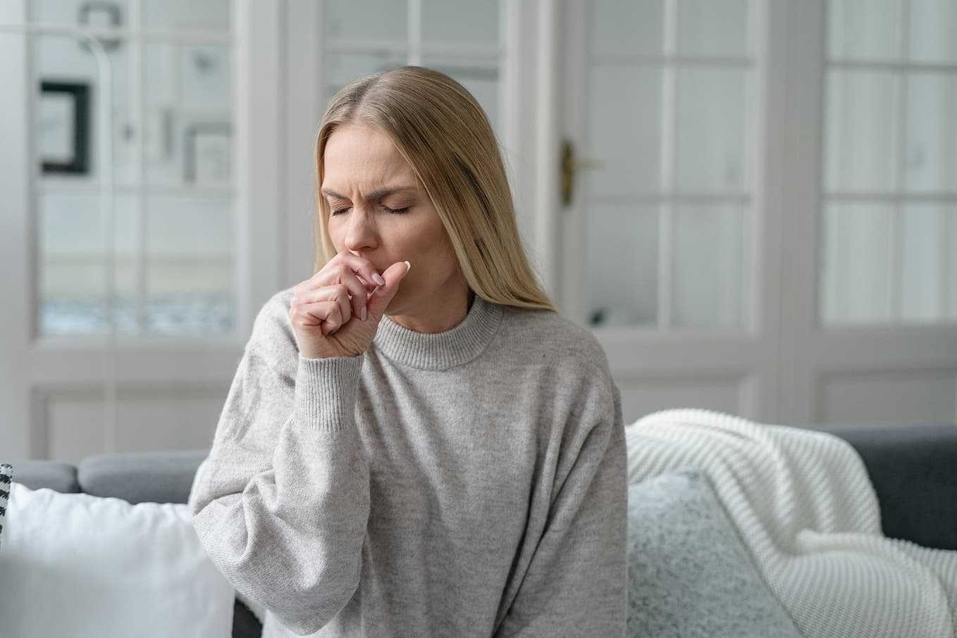 Kvinna med hosta sitter hemma i en soffa och täcker munnen med handen. Hosta kan vara ett tecken på förkylning, influensa eller luftvägsinfektion. Läs mer om orsaker, symptom och behandling.