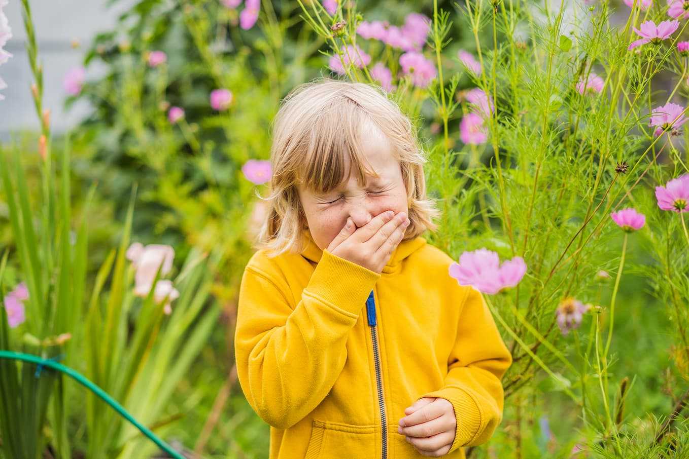 Barn med pollenallergi nyser i en blomsterträdgård. Typiska symtom på pollenallergi inkluderar nysningar, rinnande näsa och kliande ögon. Läs mer om allergisymtom och behandling på Doktera.